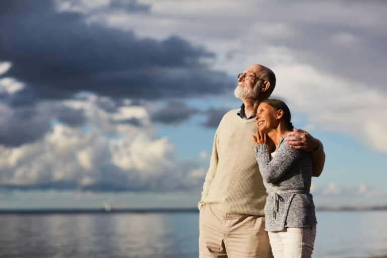 Amorous seniors standing by water against stormy sky and enjoying sundown