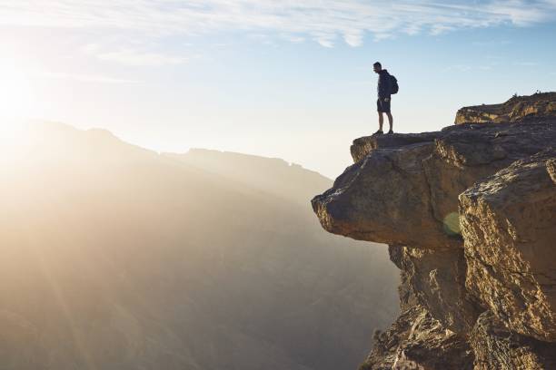 Young tourist with backpack standing on the edge of cliff at sunrise. Jebel Akhdar, Grand Canyon of Oman.